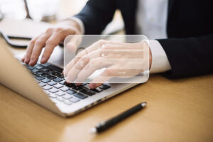 Na imagem um notebook em cima de uma mesa e mãos aparecem digitando. Em sobreposição aparece uma tela com espaço para login e senha